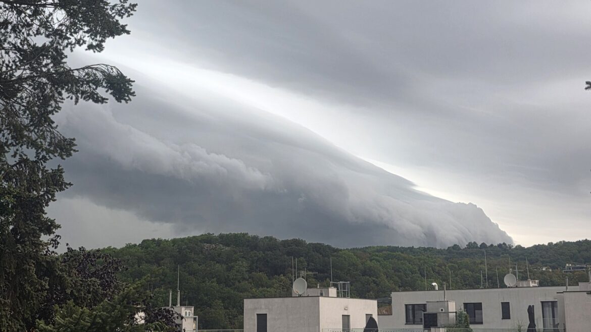 Shelf cloud, mohutný šedý oblačný val s rozbouřenou spodní stranou, se sune nad zalesněný kopec, v popředí stromy a ploché střechy domů se satelitními anténami