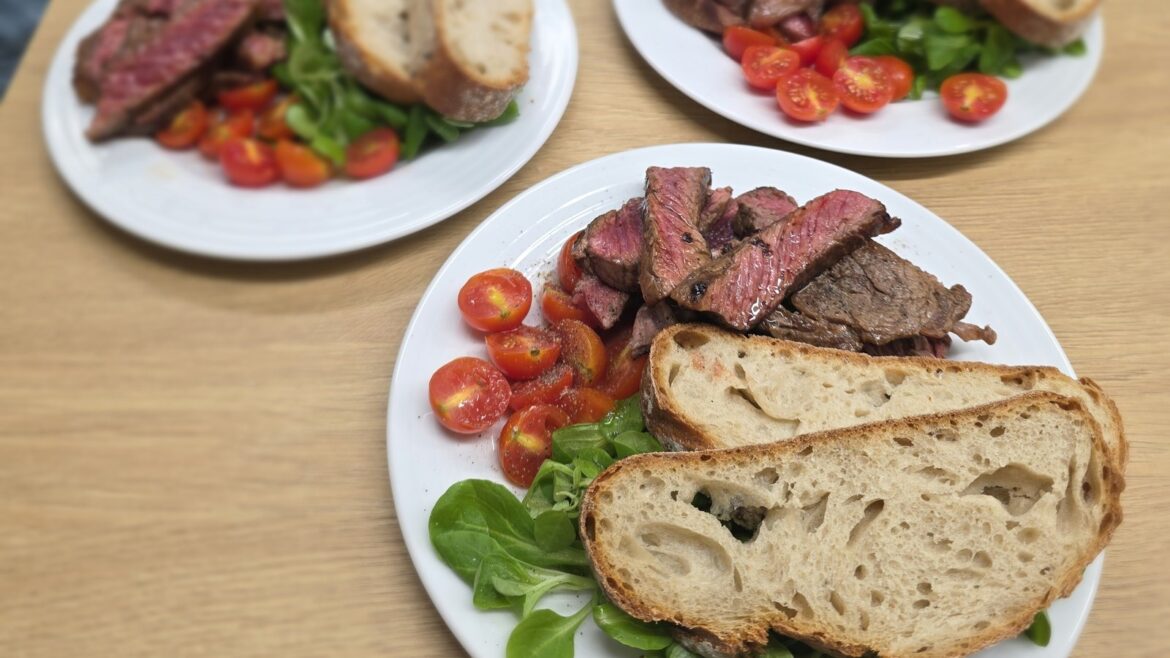 Talíř se steakem krájeným na růžové plátky s opečenou kůrkou, vedle dva krajíce kváskového chleba, půlená cherry rajčata a polníček, v pozadí další dva talíře na dřevěném stole